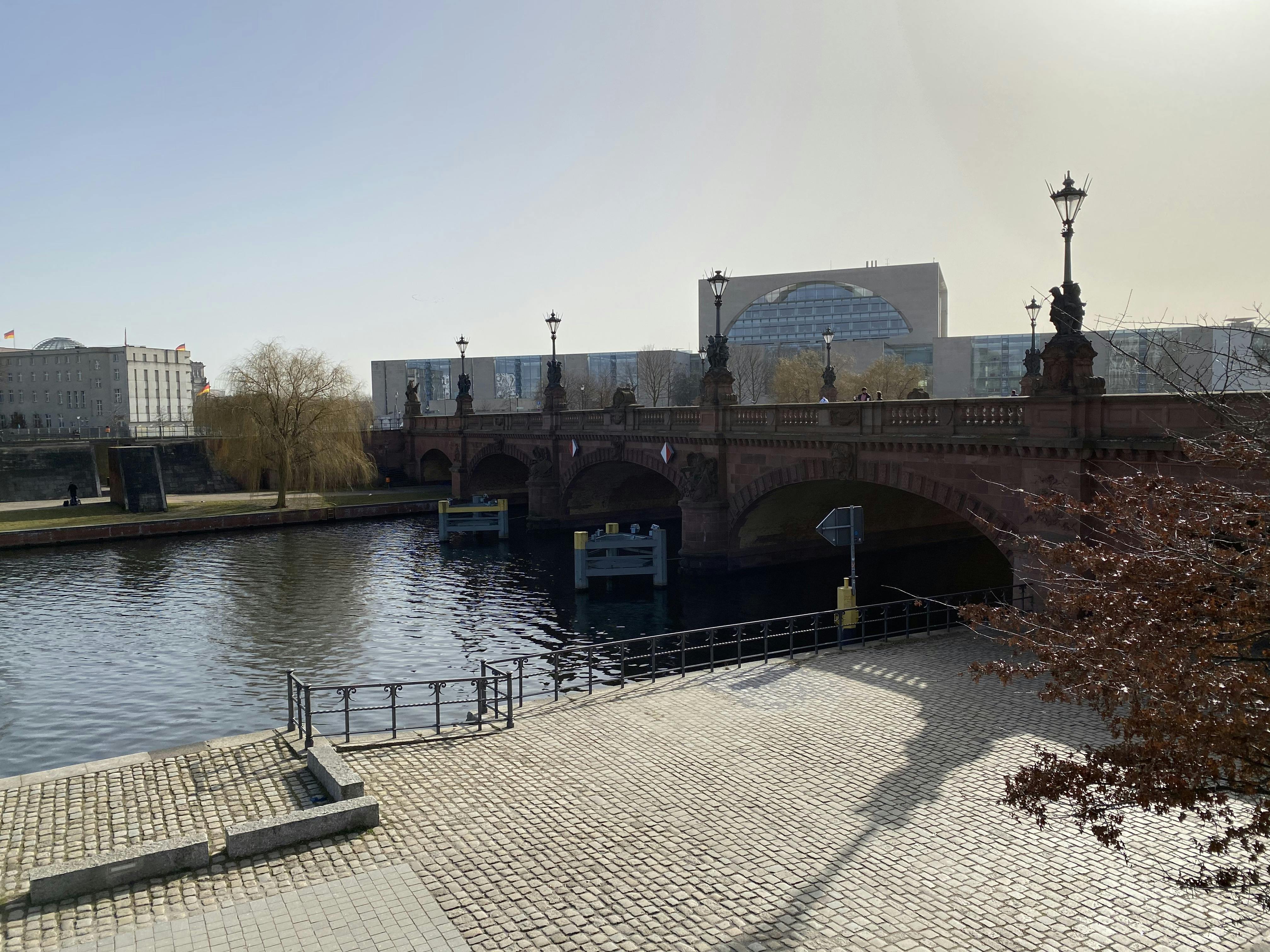 맑은 하늘 아래, 붉은 다리와 현대식 건물을 배경으로 한 강변의 자갈길.