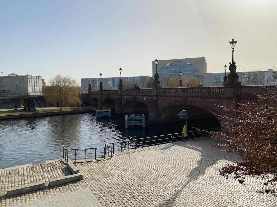 Cobblestone path along a river with a red bridge and a modern building in the background, under a clear sky.