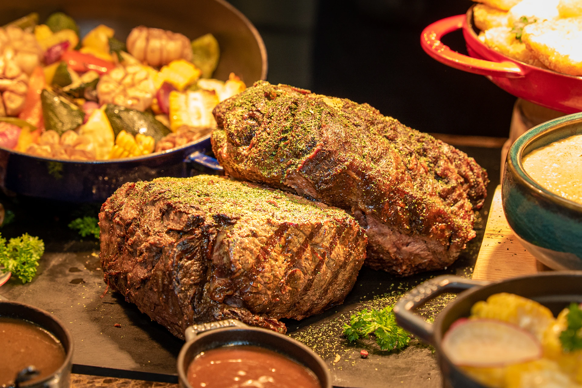 Two large, seasoned roasted meat cuts on a dark surface, garnished with herbs, with vegetables and sauce in the background.