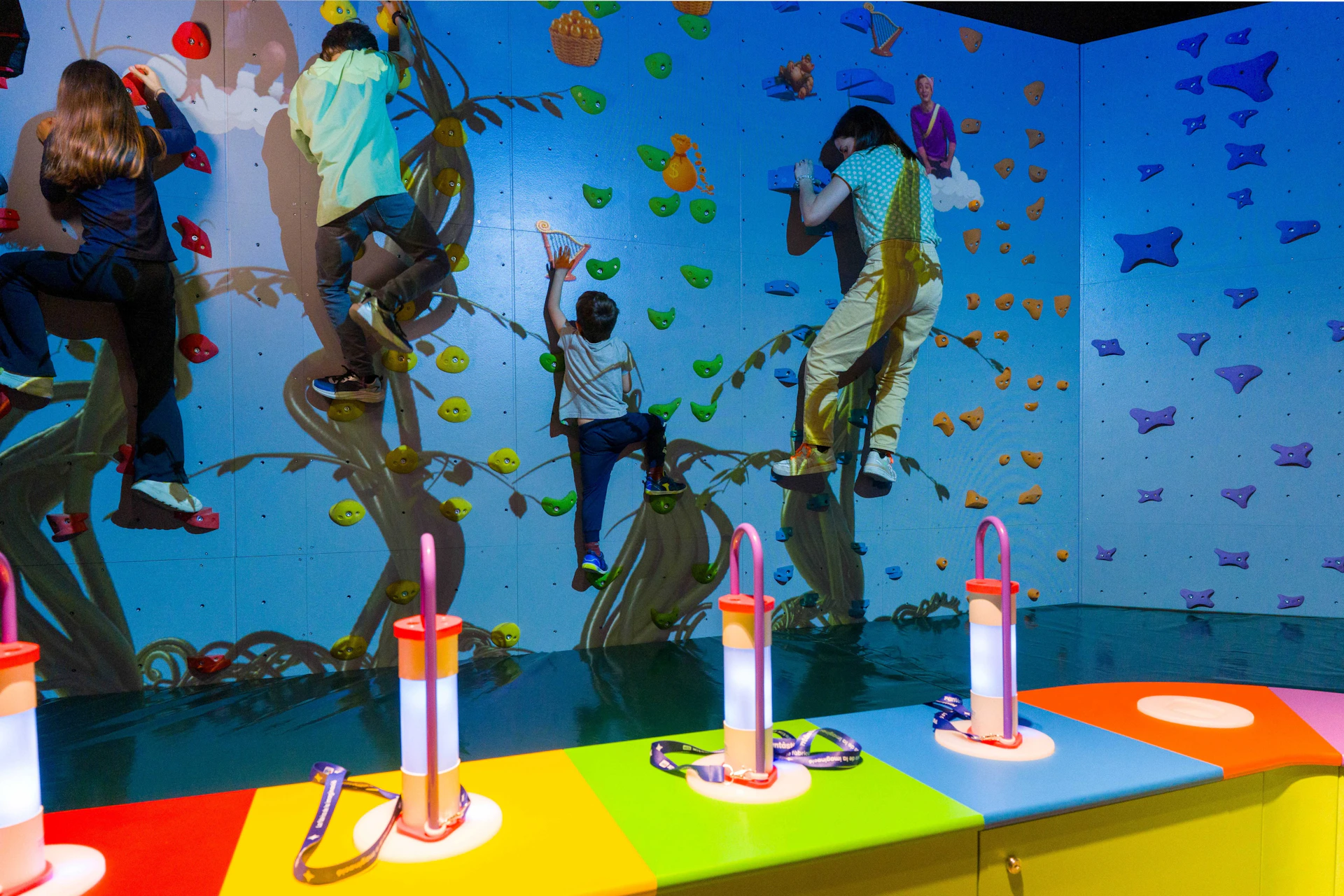 Children climbing a colorful indoor rock wall with a playful design, with illuminated equipment stands in the foreground.