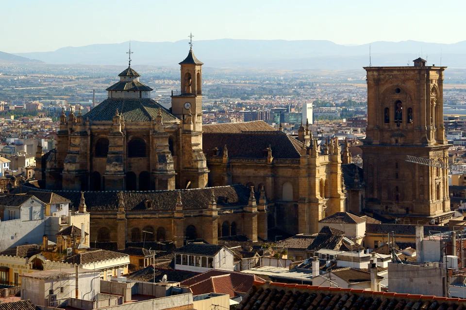 Elevated view of a historic city featuring a grand cathedral with multiple towers, surrounded by rooftops, against a mountainous backdrop.