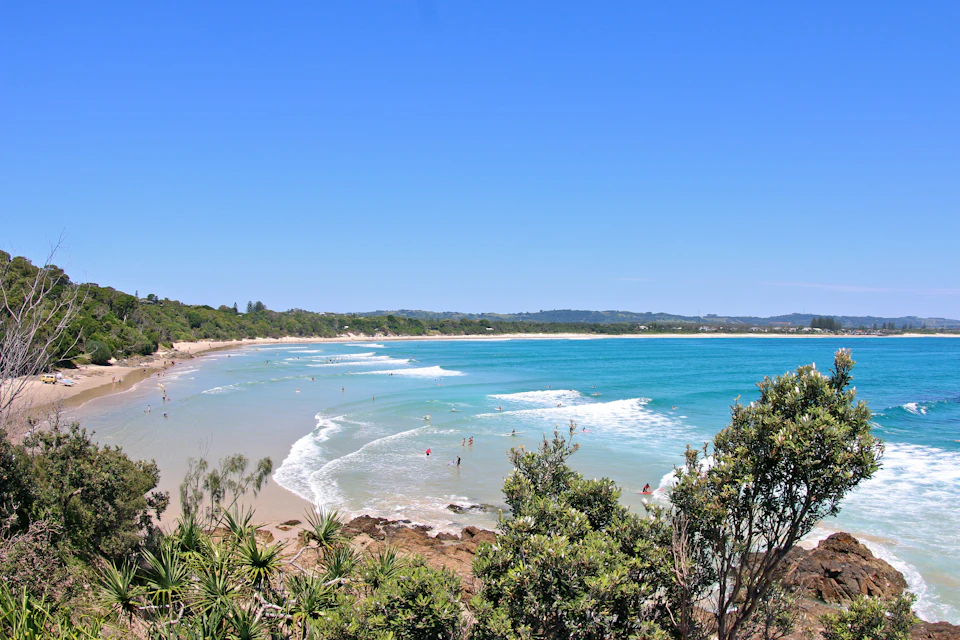 Sandy beach with gentle waves, scattered swimmers, lush greenery in the foreground, and a distant tree-lined shoreline under a clear blue sky.