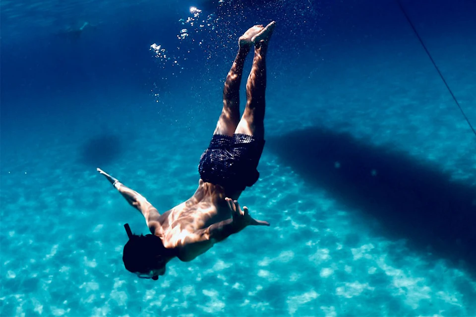 A person wearing swim trunks and a snorkel dives underwater in clear blue water with the sunlight casting shadows.