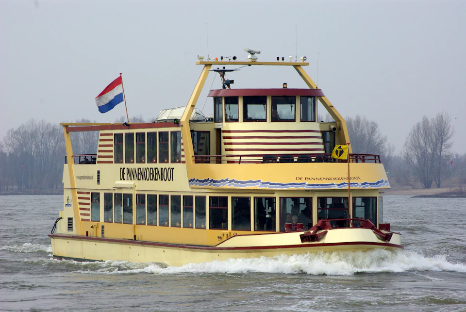 Yellow boat named "De Pannenkoekenboot" with a Dutch flag on the rear, sailing on a grey, overcast day.