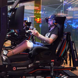 En person i en spelstol med racing-simulator, som bär ett headset och fokuserar på skärmen, inomhus med färgstark belysning.