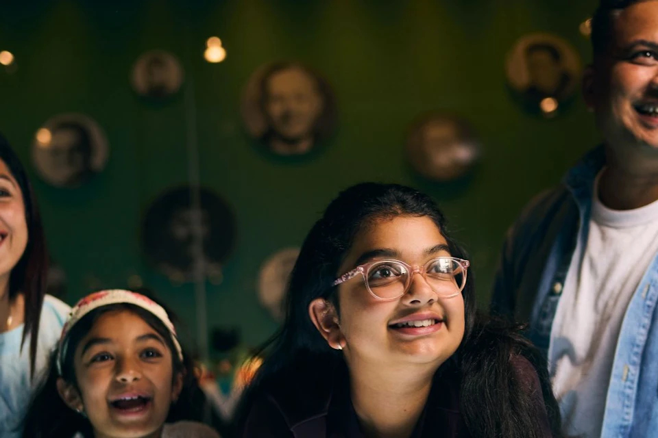 A family of four interacts with an illuminated display in a museum-like setting, smiling and engaged.