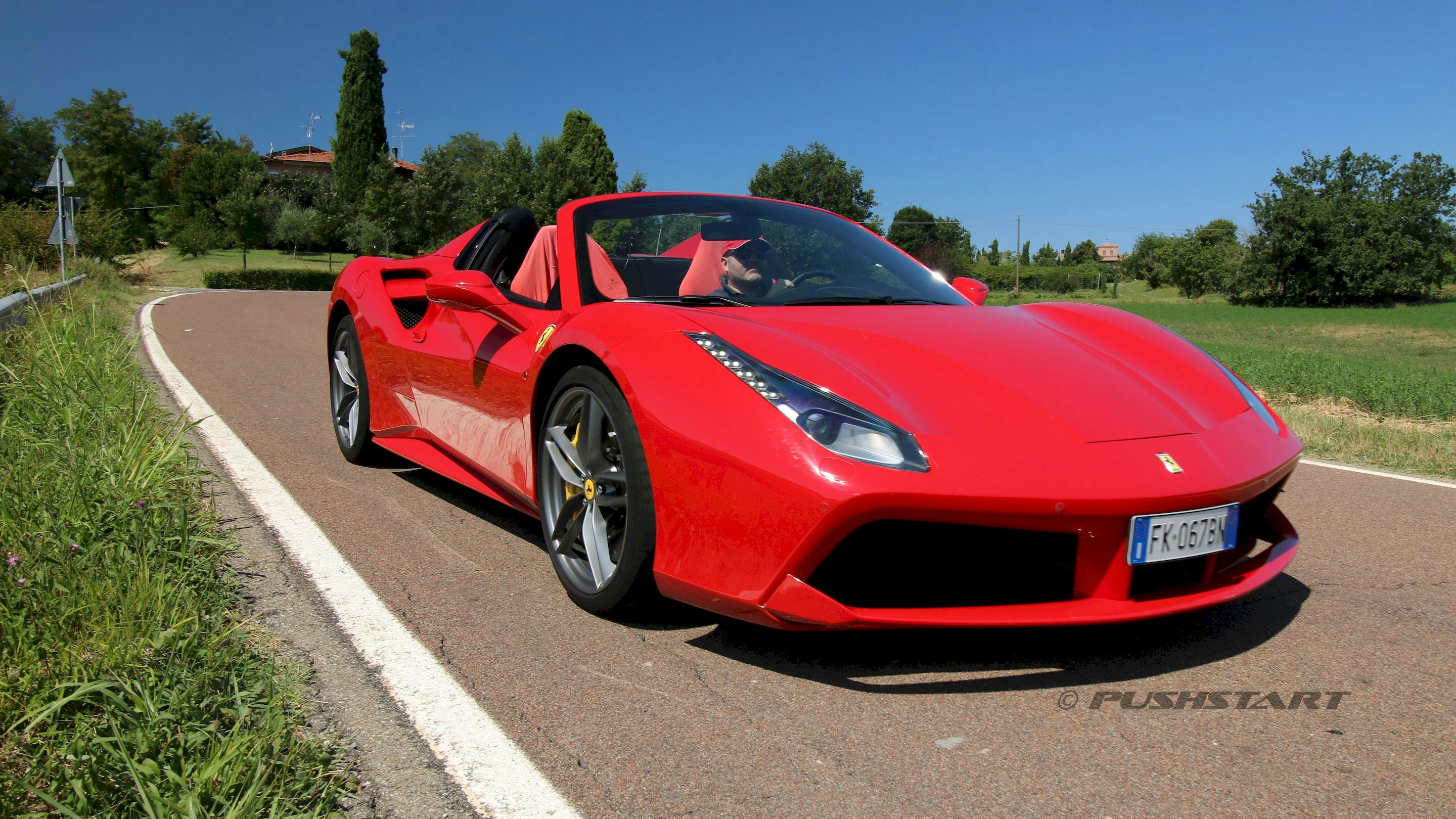 Red sports car with a man driving on a sunny road, surrounded by greenery and trees in the background.