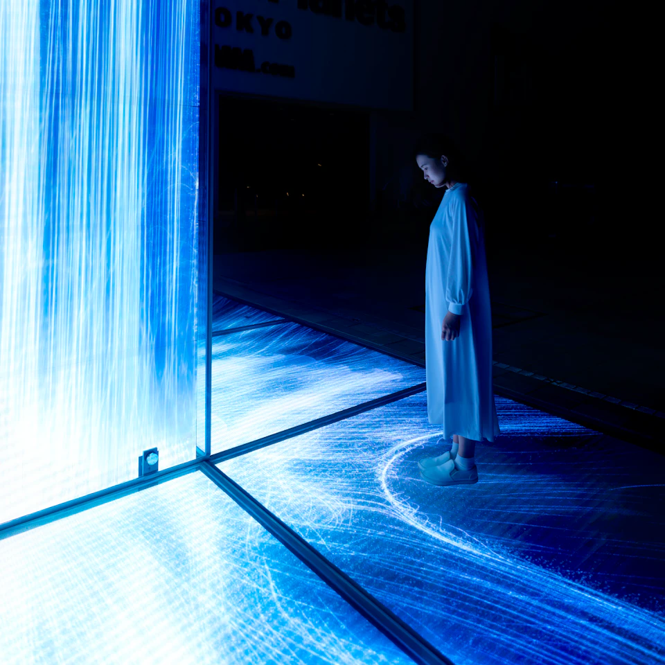 A person in a white dress stands in a dark room next to an illuminated floor and wall displaying flowing blue and purple light patterns.
