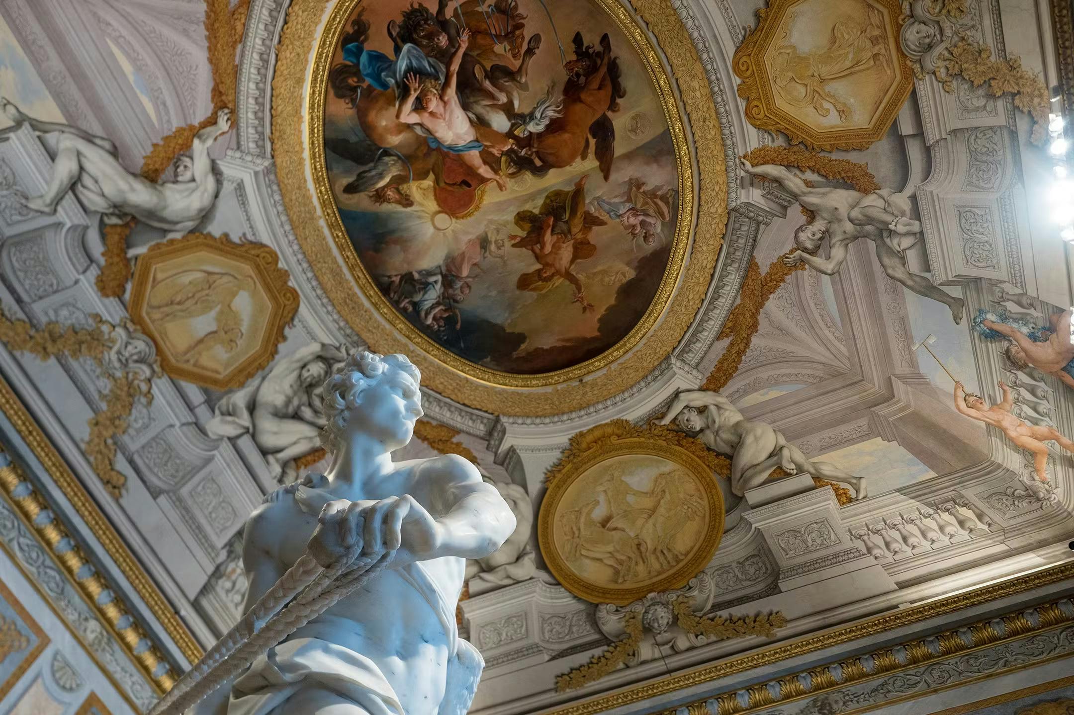 Ornate room with a ceiling fresco of mythological figures and a white marble statue in the foreground, surrounded by intricate carvings.