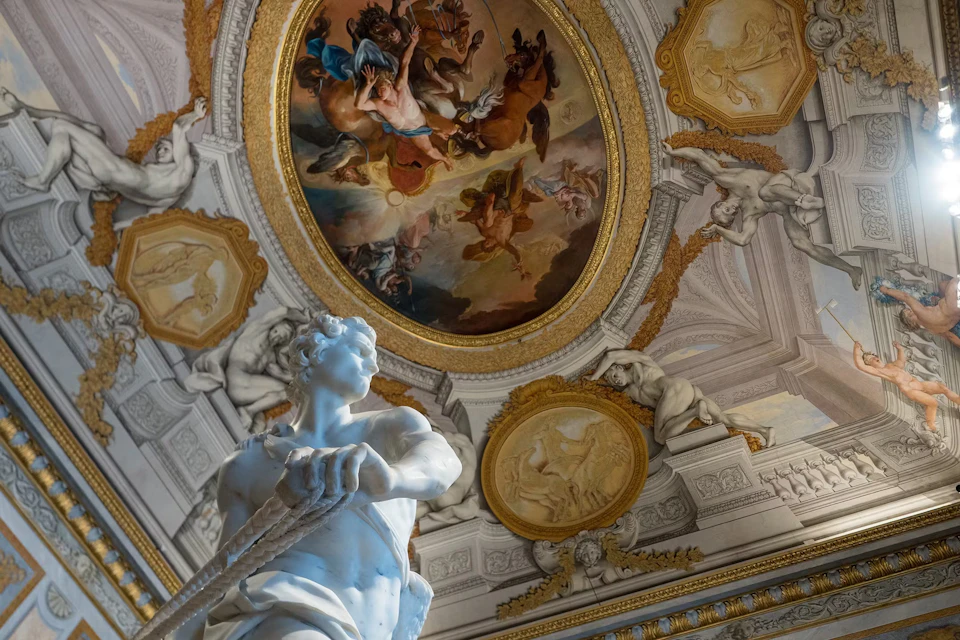 Ornate room with a ceiling fresco of mythological figures and a white marble statue in the foreground, surrounded by intricate carvings.