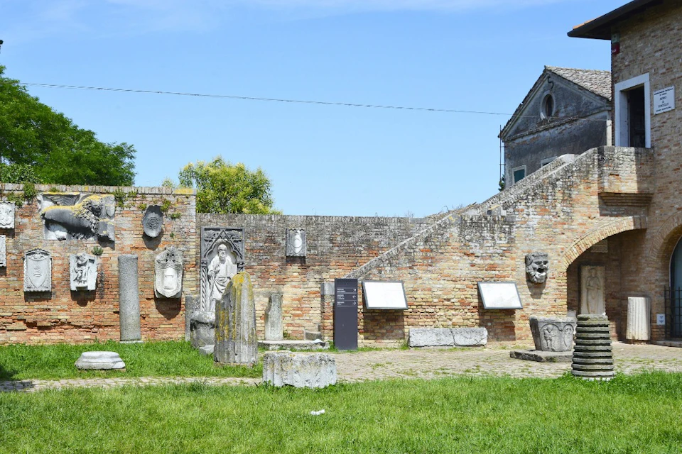 Outdoor museum exhibit with stone artifacts and sculptures displayed against a brick wall, with grass in the foreground.