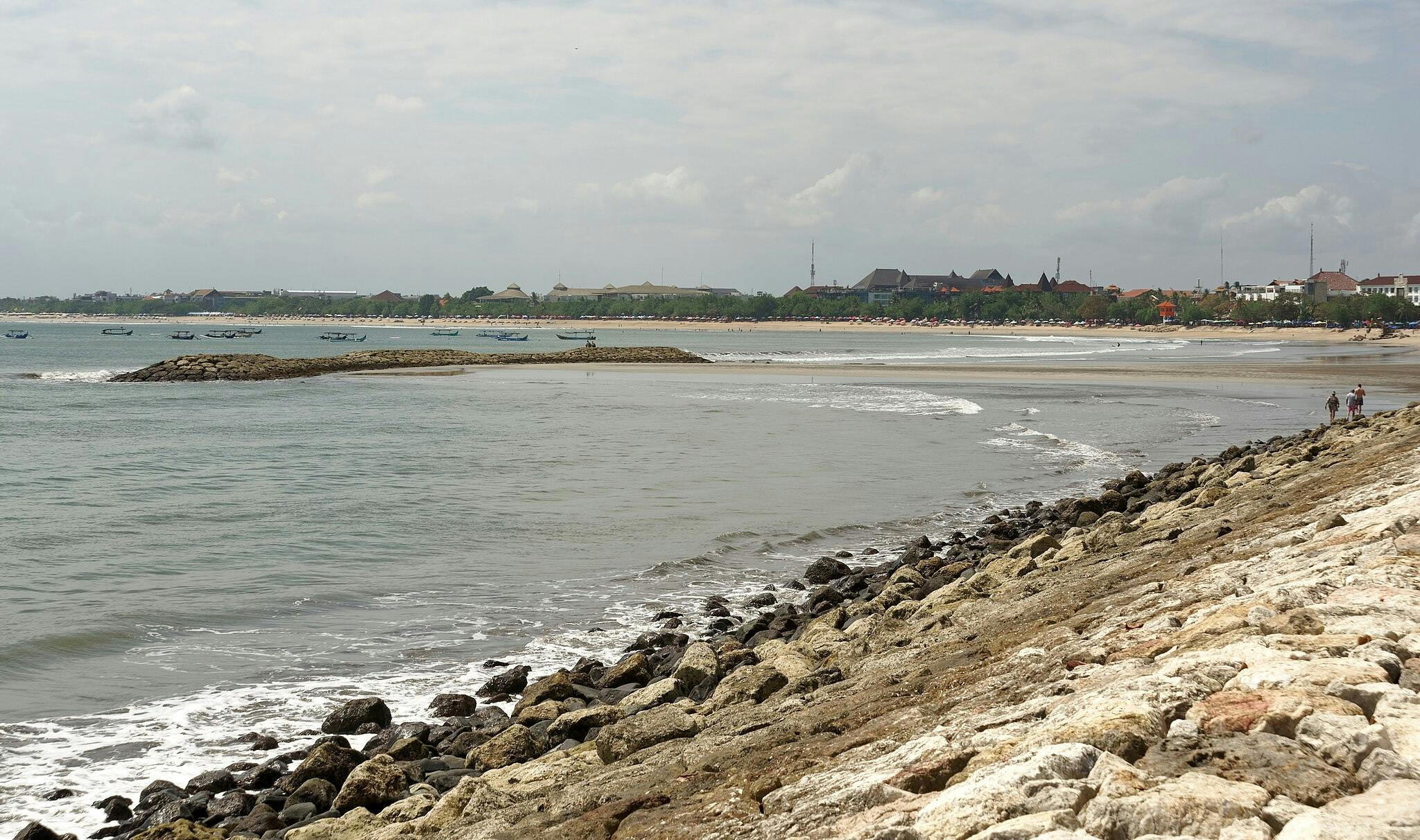 Stenig strandlinje som leder till en sandstrand med båtar i vattnet och byggnader i fjärran under en molnig himmel. Två personer går längs klipporna.