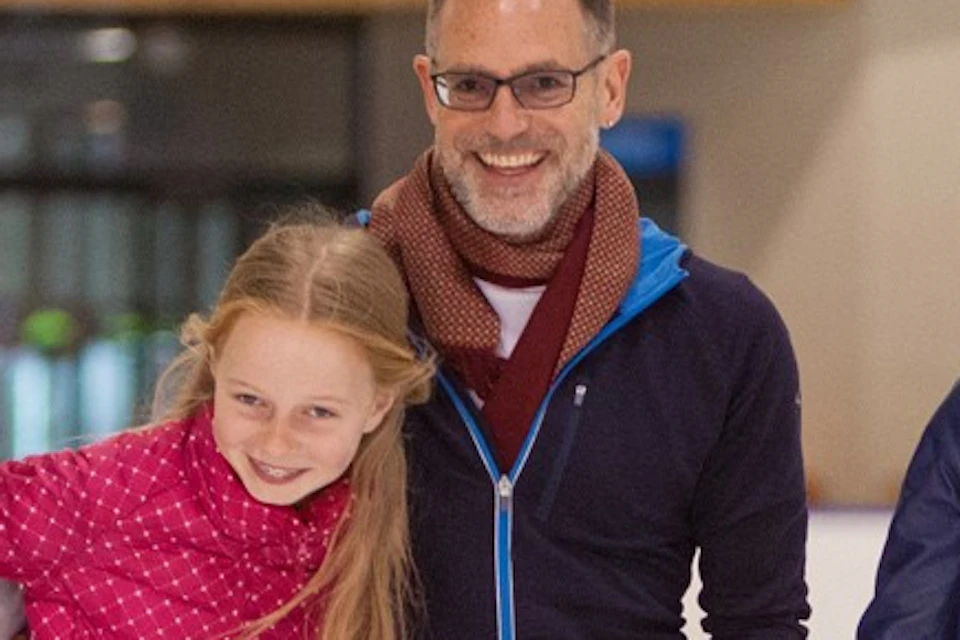 A family of four skating together on an indoor ice rink, smiling and holding hands.