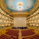 Ornate theater with red seats, a large stage, and elaborate gold-trimmed balconies under a painted, chandelier-adorned ceiling.