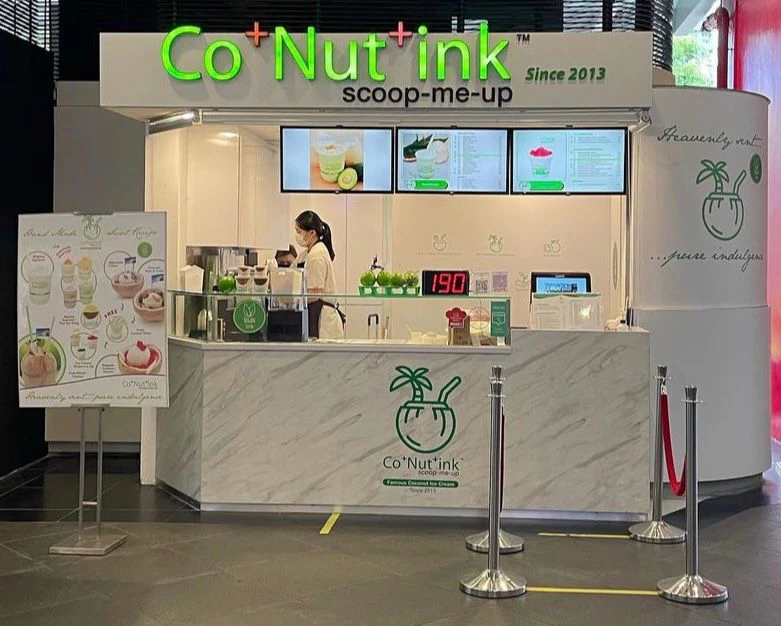 A Co+Nut+ink dessert kiosk with a staff member preparing an item. A menu display and a poster are visible.