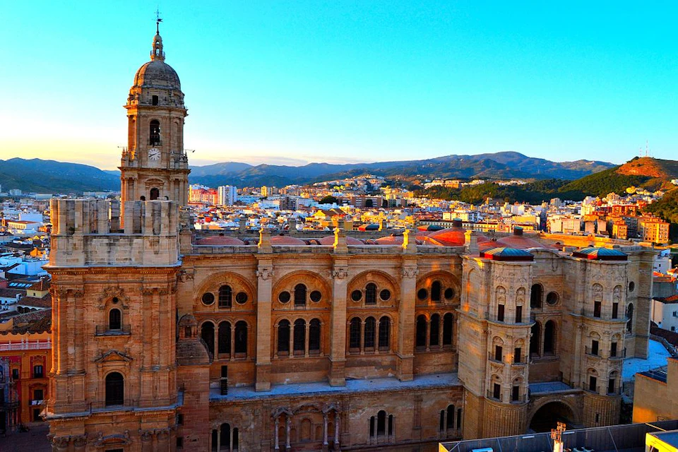 Historic cathedral with arched windows and a tall clock tower against a cityscape and mountains under a clear blue sky.