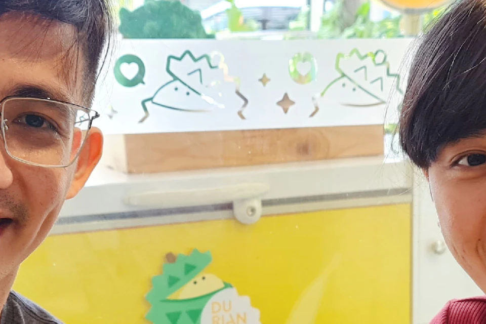 Two people smiling and holding trays with durian pieces labeled with tags, in a bright indoor setting.