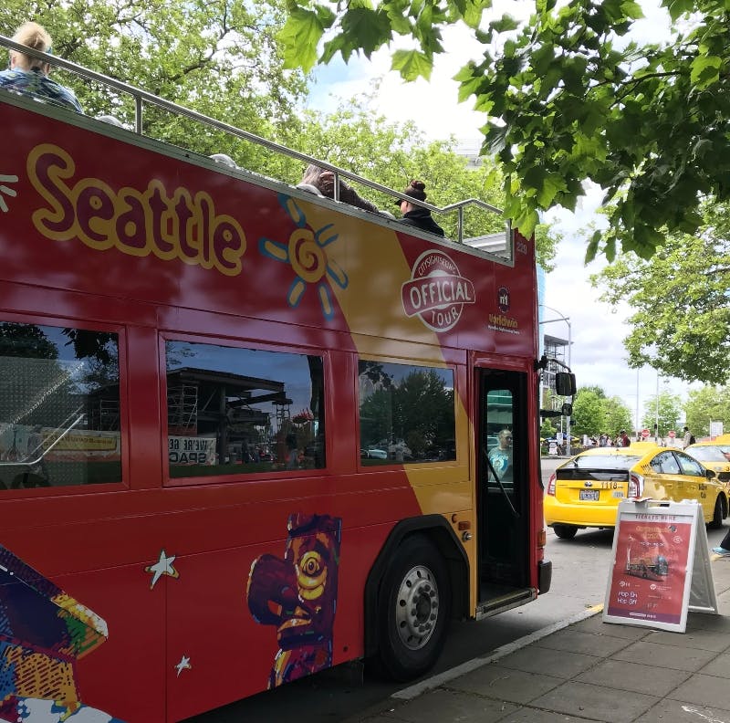 Red double-decker tour bus parked on a tree-lined street with "Seattle" written on the side. A taxi and a tour sign are nearby.
