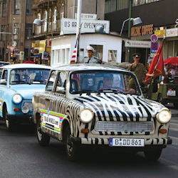 En rad färgglatt målade veteranbilar kör förbi en replik av den amerikanska arméns checkpoint i en urban miljö. Vakterna står redo.