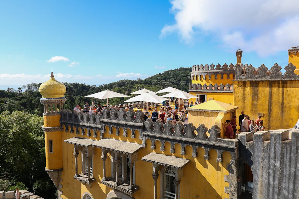 A yellow castle with gothic architecture, a terrace full of people under umbrellas, and surrounded by greenery.