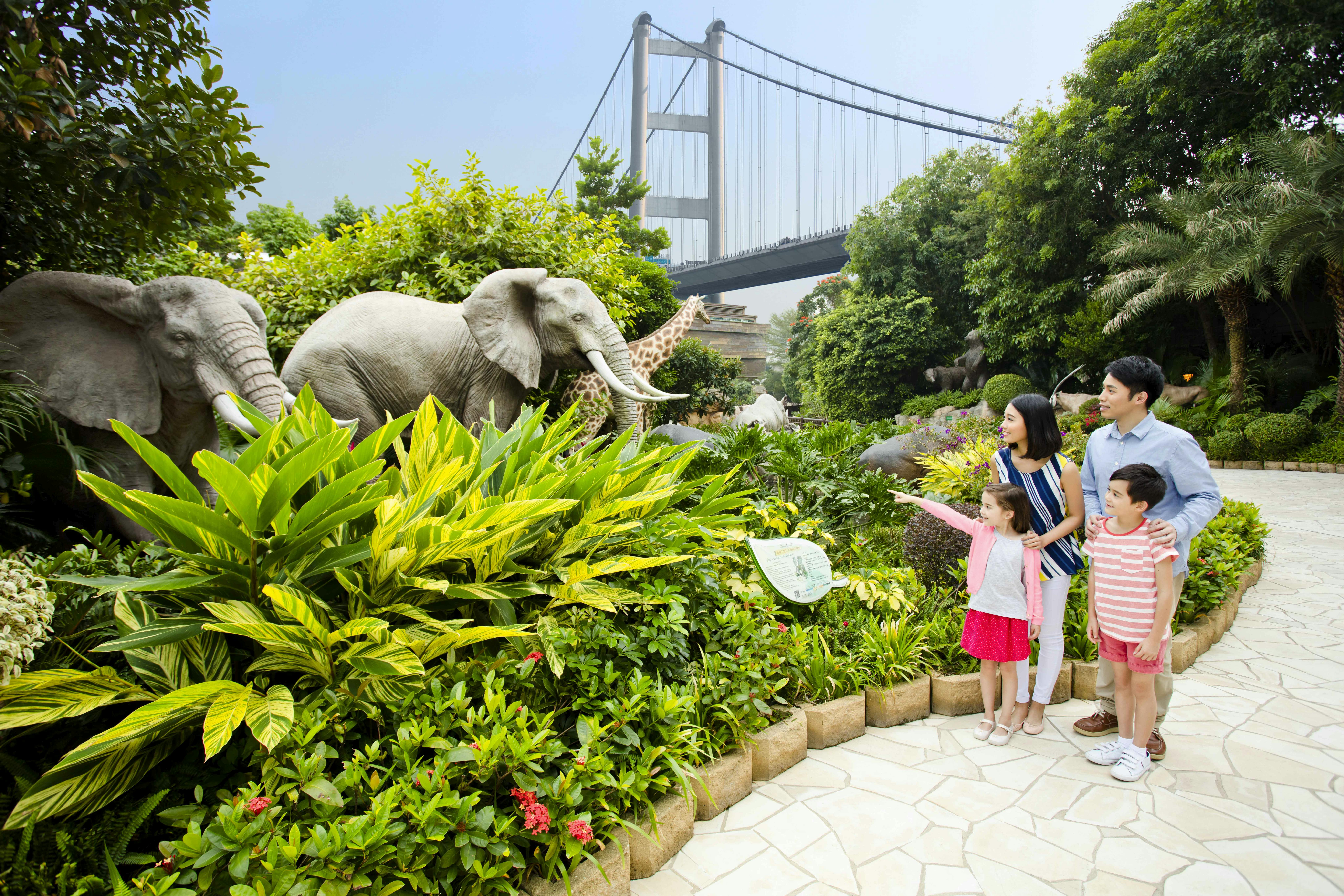 Семья из четырех человек любуется статуями животных в пышном парке с мостом на заднем плане.