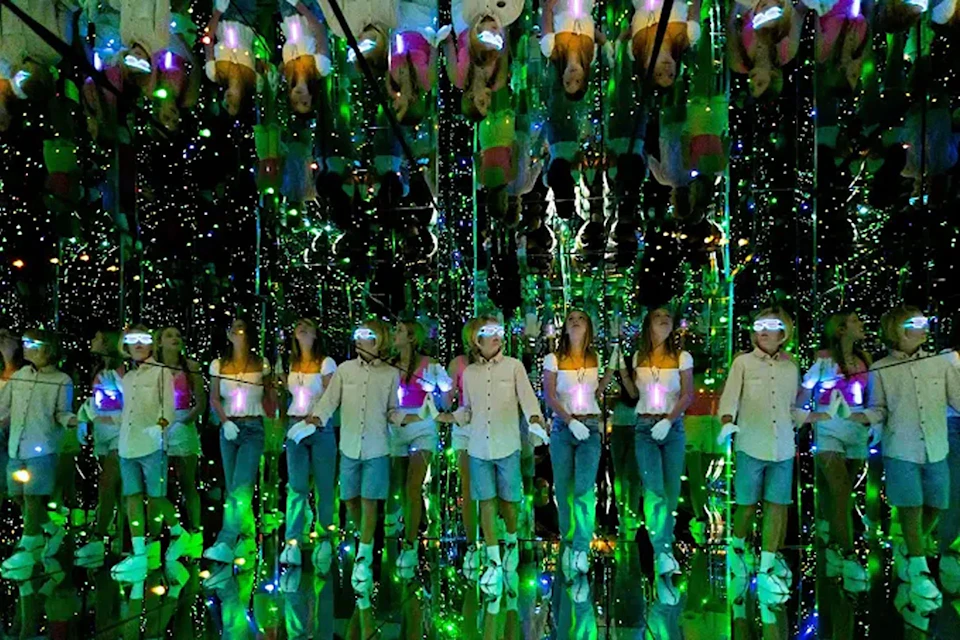 A group of people in a mirrored room with green and other colored lights, creating multiple reflections.
