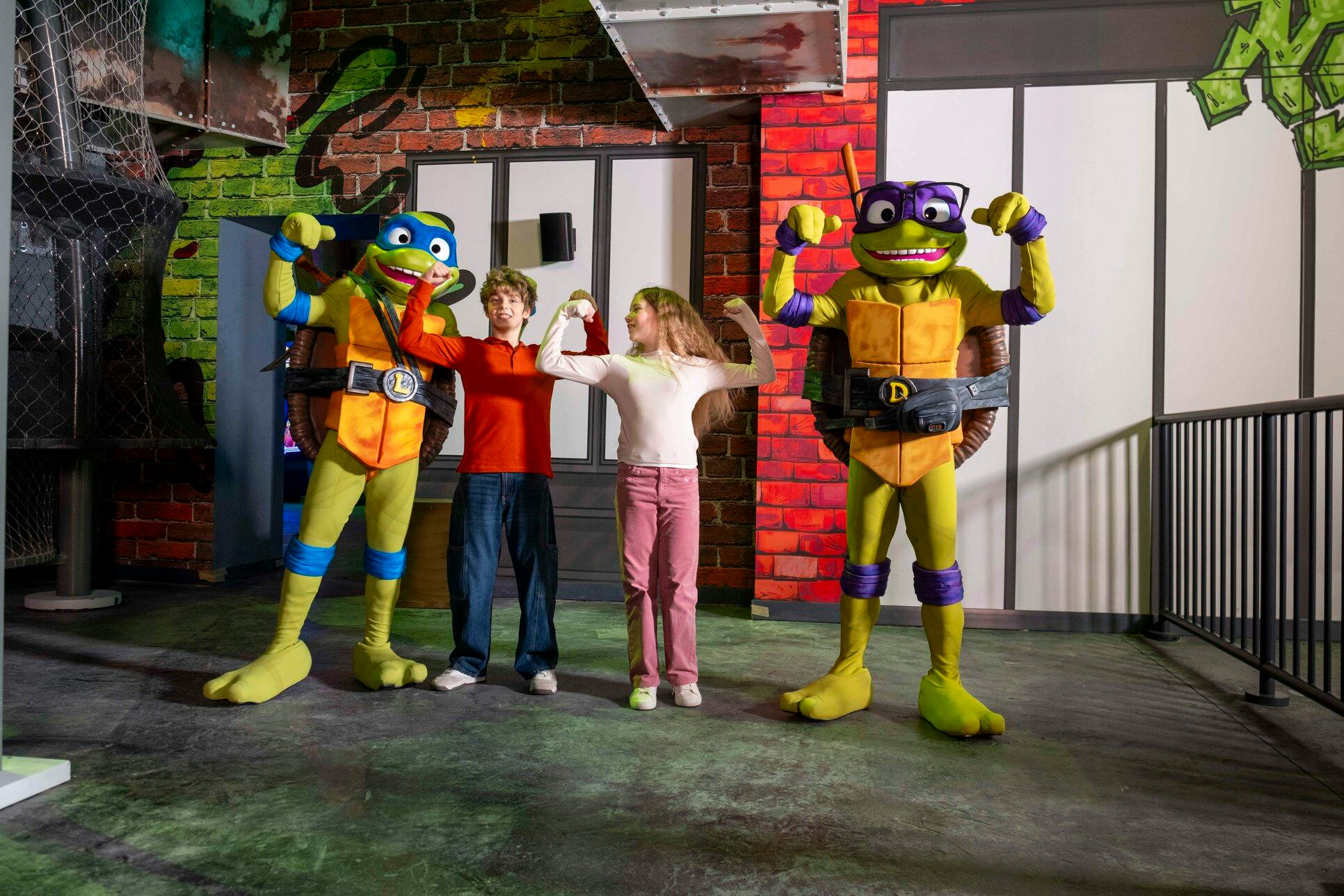 Twee kinderen poseren met gekostumeerde Ninja Turtle-figuren in een kleurrijke binnenomgeving.