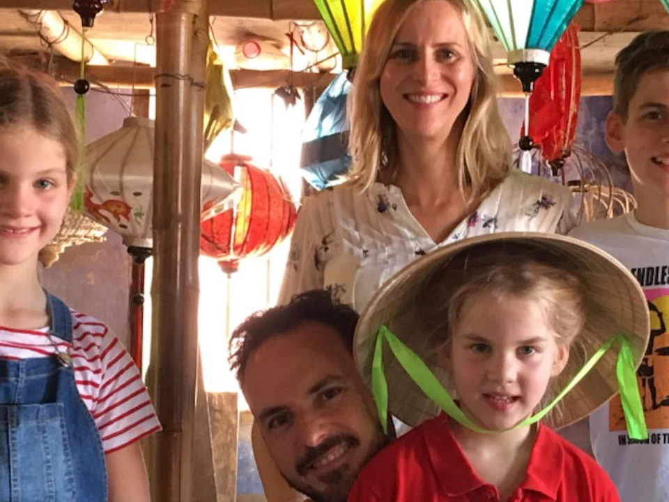 A family of five smiling under colorful hanging lanterns in a rustic setting, with wooden poles and baskets visible.