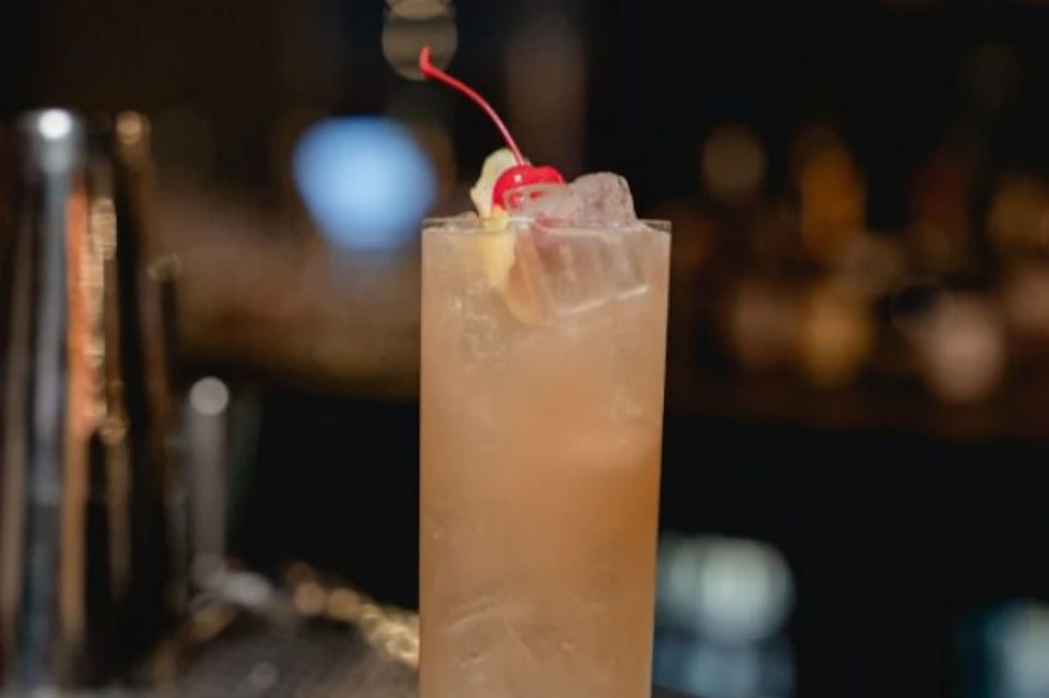 A tall glass filled with a light brown drink, ice, a cherry, and a lemon wedge garnish, set against a dimly lit background.