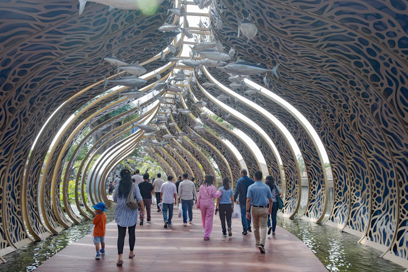 People walking through a decorative, fish-themed arched walkway over water, surrounded by intricate metallic patterns.
