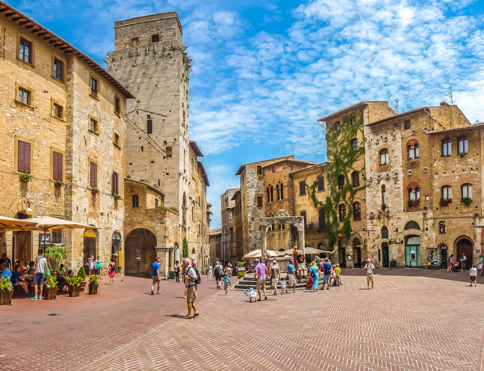 People walk and gather in a historic town square with stone buildings, a tower, and outdoor cafes under a bright blue sky.