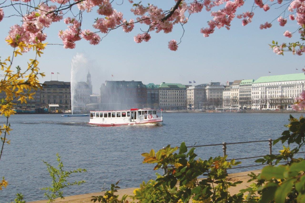 Una barca bianca e rossa su un lago con una fontana sullo sfondo, incorniciata da fiori rosa ed edifici cittadini.
