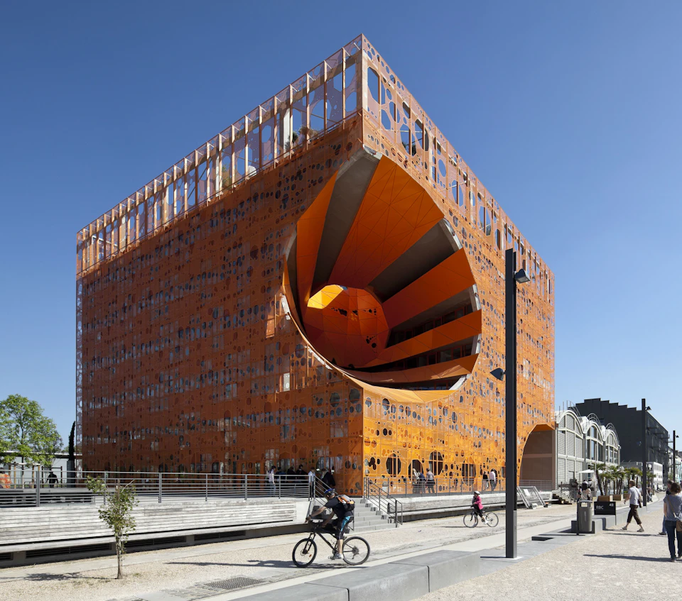 Modern orange cubic building with a large circular cutout on one side. People walk and bike in the foreground under a clear blue sky.