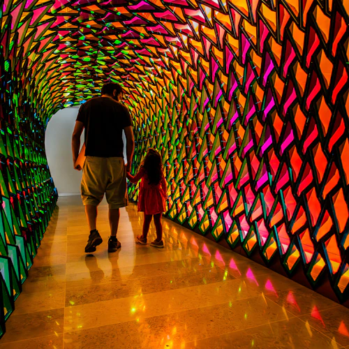 En man och ett barn går genom en färgglad, geometriskt mönstrad tunnel med levande ljus som reflekteras på golvet.