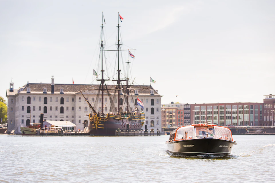 A canal with a modern boat in the foreground and a large, historic sailing ship docked near buildings in the background.