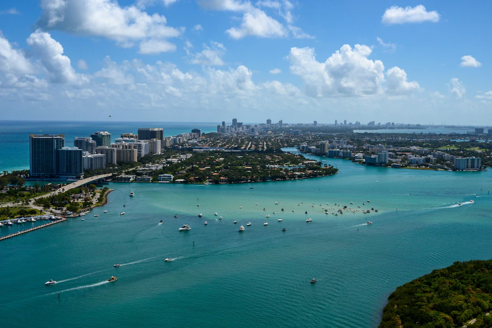 A coastal cityscape with buildings, waterways, and numerous boats in the water under a partly cloudy sky.