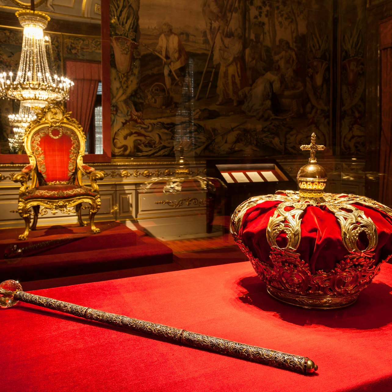 En gyllene spira och utsmyckad krona sitter på röd sammet i ett stort rum med en förgylld stol och ljuskrona.