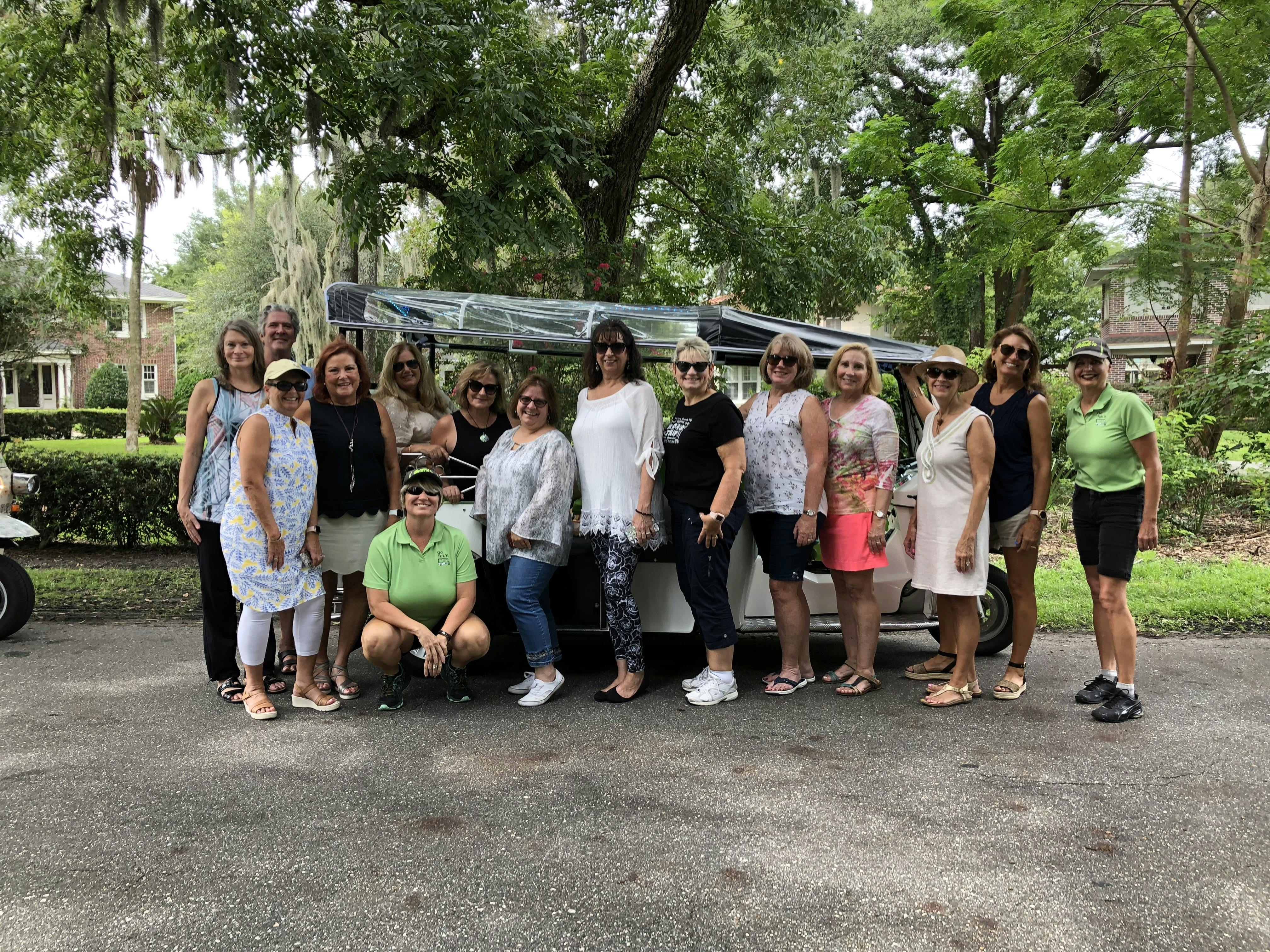 En gruppe på 14 personer poserer sammen foran et hvidt køretøj i et frodigt nabolag med træer og murstenshuse.