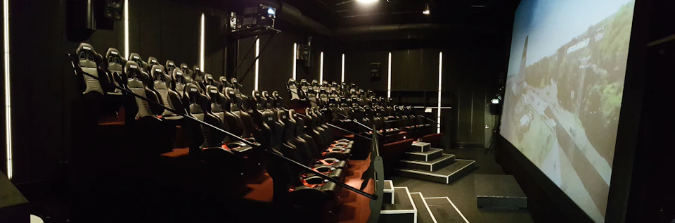 A dimly lit theater with multiple rows of black and red motion seats, facing a projector and a screen area.