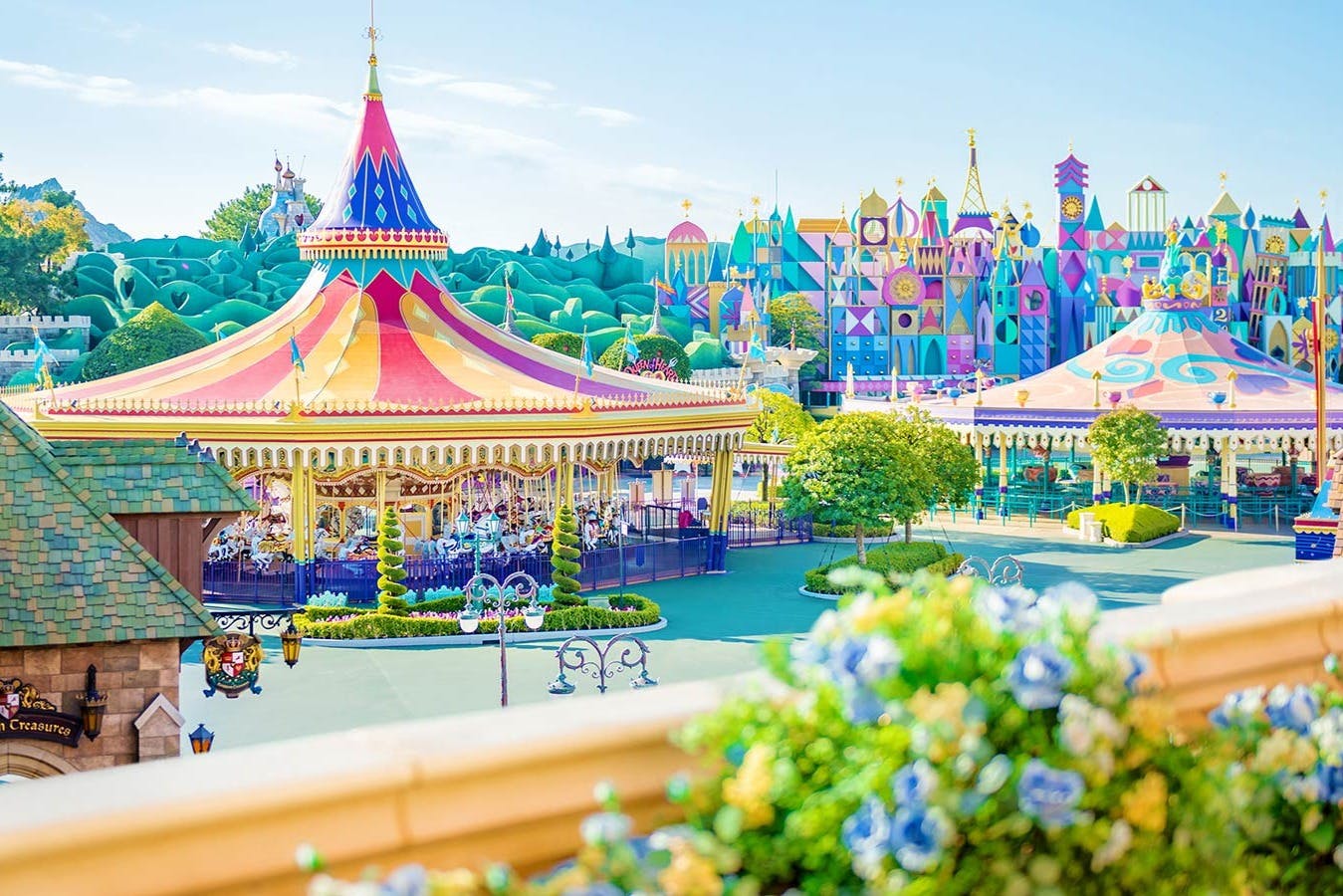 Έντονα χρωματισμένο λούνα παρκ με ένα καρουζέλ κάτω από μια κόκκινη και κίτρινη σκηνή, ιδιότροπα κτίρια και καταπράσινο τοπίο σε μια ηλιόλουστη μέρα.