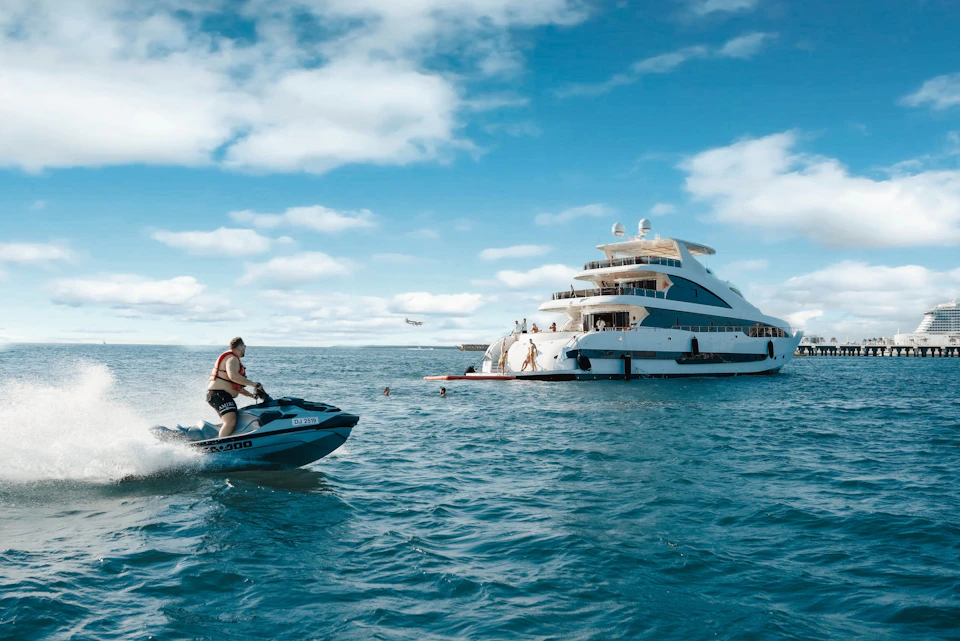 A person rides a jet ski near a large yacht on a sunny day with a few other people on the yacht's deck.