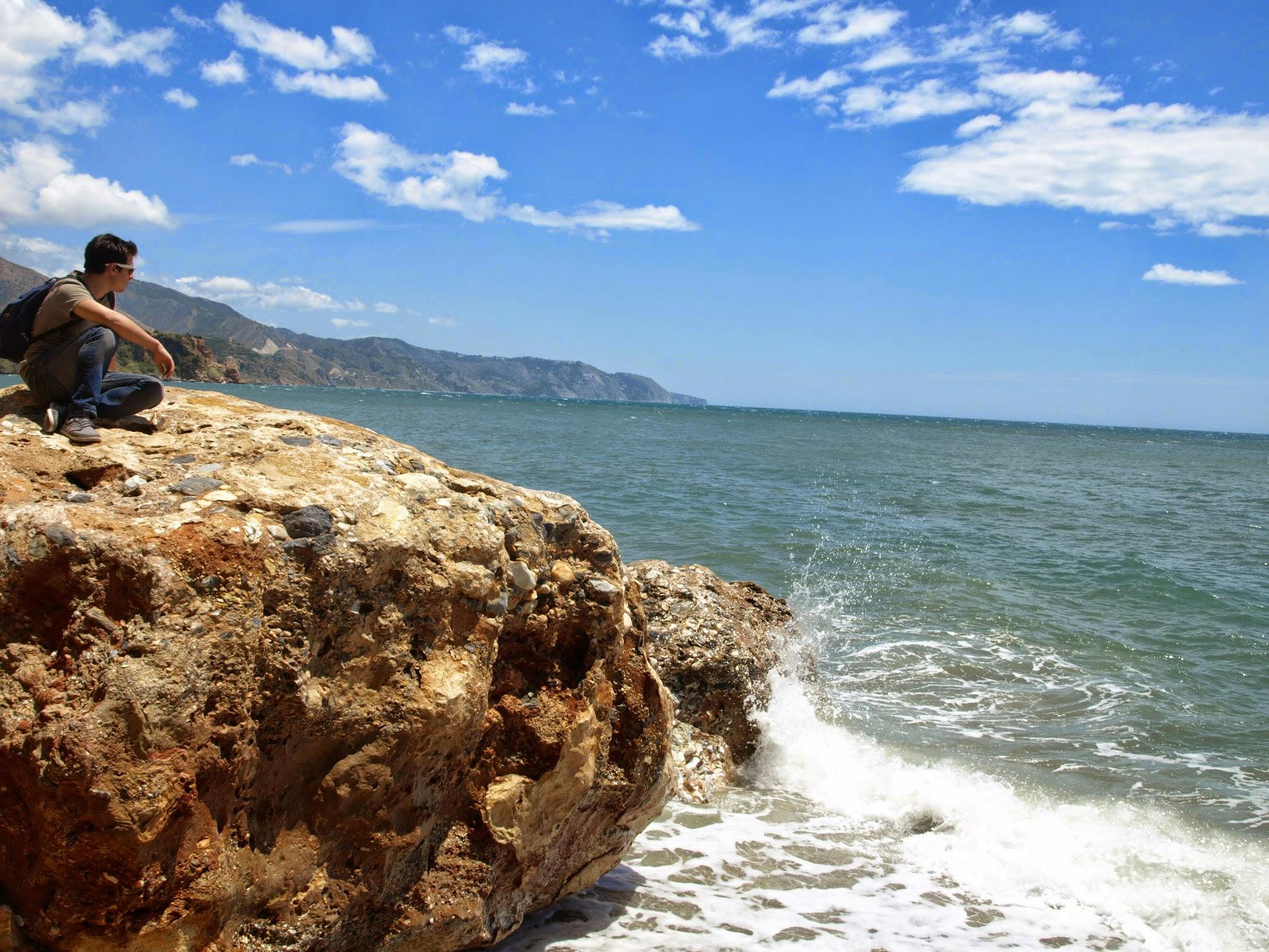 Person som sitter på stora stenar vid havet med vågor som kraschar. Klar himmel med en del moln och avlägsna berg är synliga.