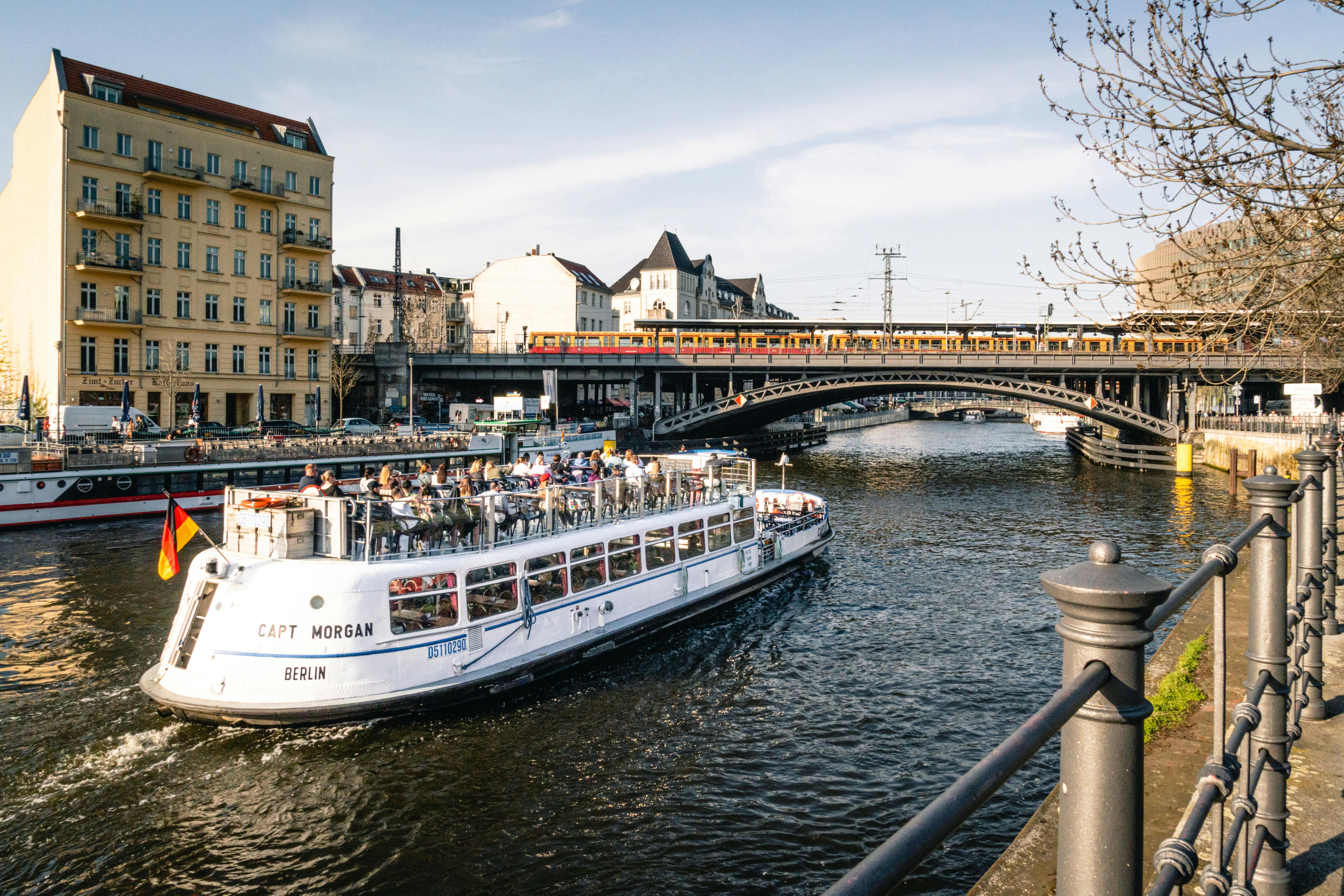 Un bateau d'excursion avec des passagers navigue sur une rivière sous un pont où passe un train, entouré de bâtiments urbains.