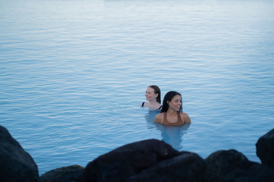 Two people in a calm, blue body of water, with dark rocks in the foreground.
