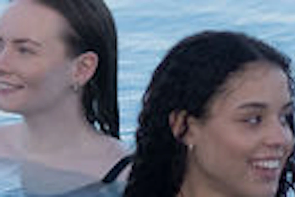 Two people in a calm, blue body of water, with dark rocks in the foreground.