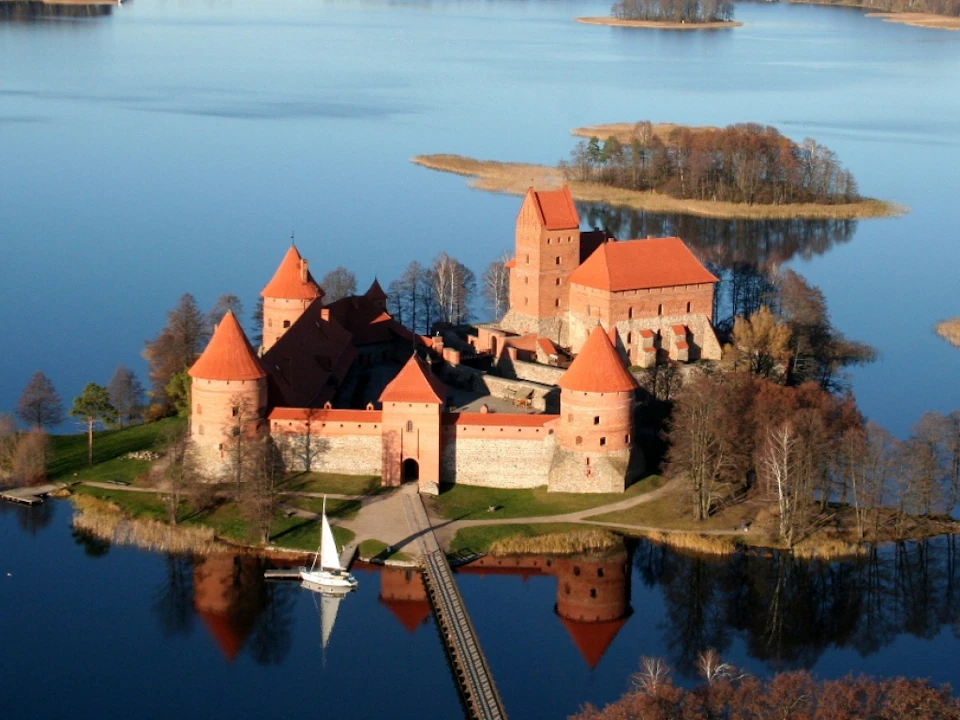 Aerial view of a castle with red-roofed towers surrounded by a lake, connected to land by a wooden bridge and a sailboat nearby.