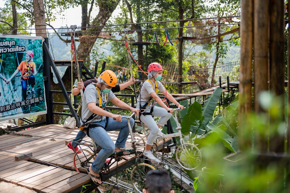 People in safety gear riding bicycles on a zipline in a forested area, with harnesses and helmets for protection.