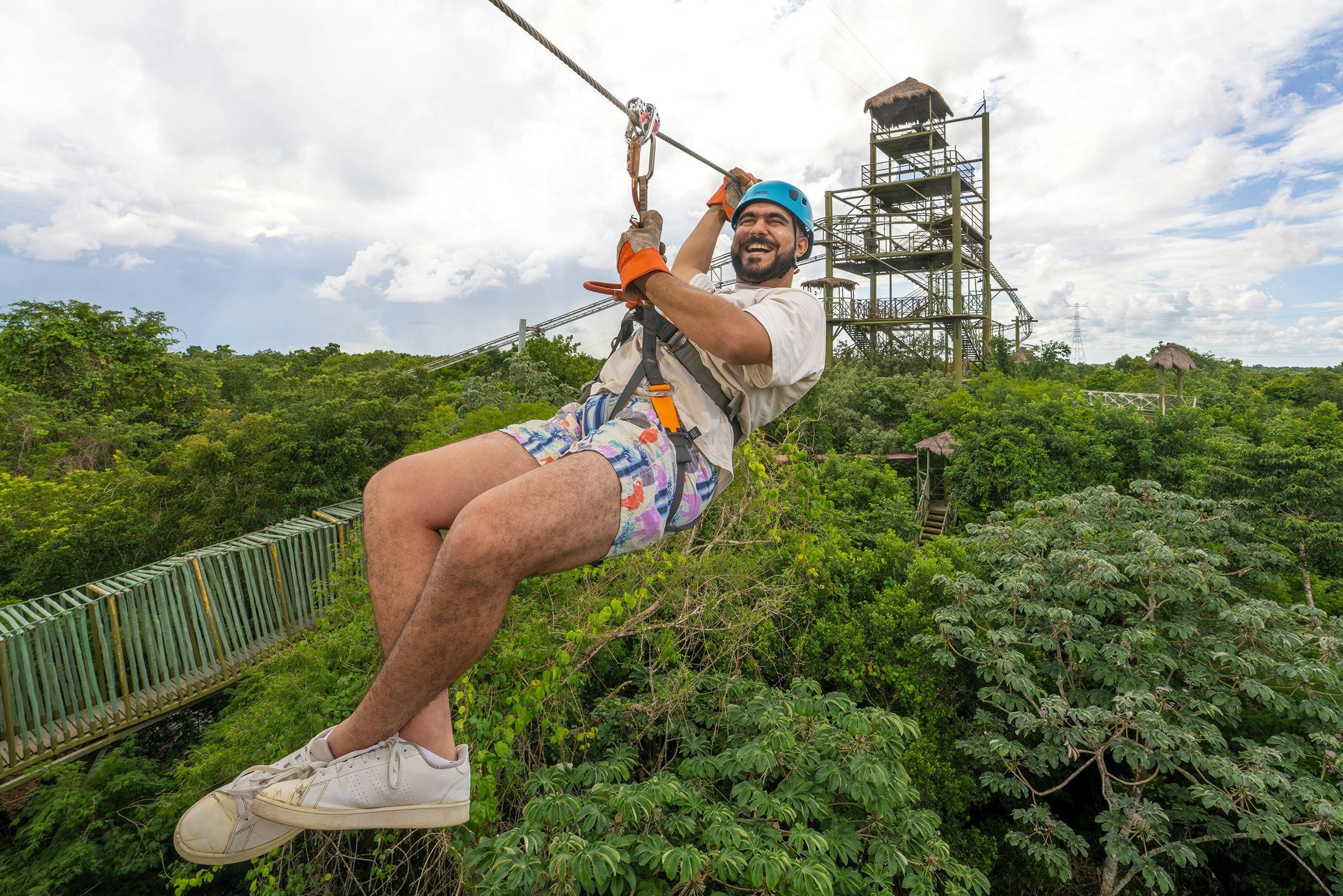 青いヘルメットとカラフルなショートパンツを着た人が、緑豊かな緑の上のジップラインに乗っています。背景には高層の展望タワーがあります。