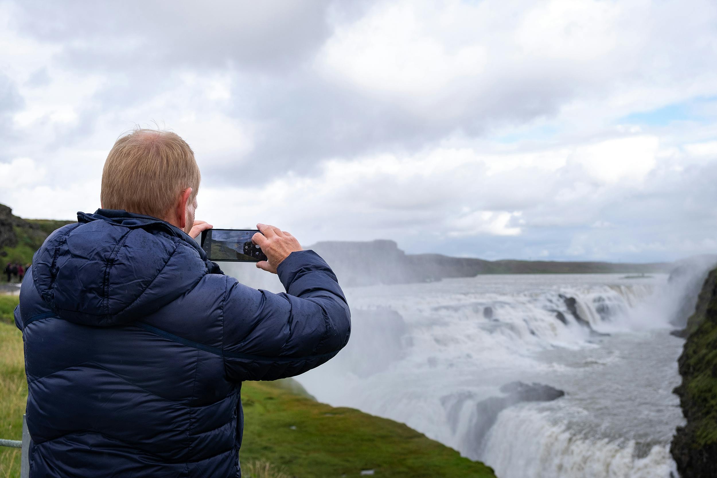 A person in a blue jacket photographs a large waterfall under a cloudy sky.
