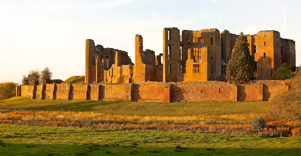 Ruins of a large, orange-brick building stand on a grassy field under a clear blue sky.